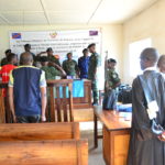 The inside of the Tribunal de Paix de Kalehe. Soldiers stand around a desk at the front of the room, judges stand at a desk on the side of the room, and people stand behind a guard rail. A banner in French covers the wall at the front of the room.