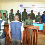 The inside of the Tribunal de Paix de Kalehe. Soldiers stand around a desk at the front of the room and three people stand behind a guard rail. A banner in French covers the wall at the front of the room.