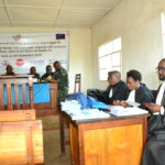 The inside of the Tribunal de Paix de Kalehe. Soldiers sit around a desk at the front of the room and three judges sit behind a desk on the side of the room. A banner in French covers the wall at the front of the room.