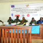 The inside of the Tribunal de Paix de Kalehe. Soldiers sit around a desk at the front of the room. A banner in French covers the wall at the front of the room.