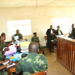 The inside of the Tribunal de Paix de Kalehe. Soldiers sit around a desk at the front of the room looking through papers, and three judges stand at a desk on the side of the room. A banner in French covers the wall at the front of the room.