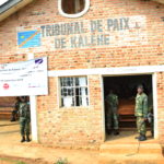 The outside of the building for the Tribunal de Paix de Kalehe. Guards stand in the entryways, some hold guns. A banner hangs over one of the entryways.